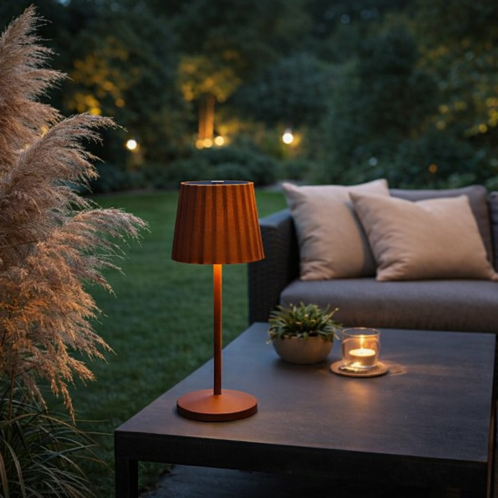 Eine braune LED-Tischleuchten mit geriffeltem Lampenschirm auf einem Tisch im Garten neben einer Sofaecke.