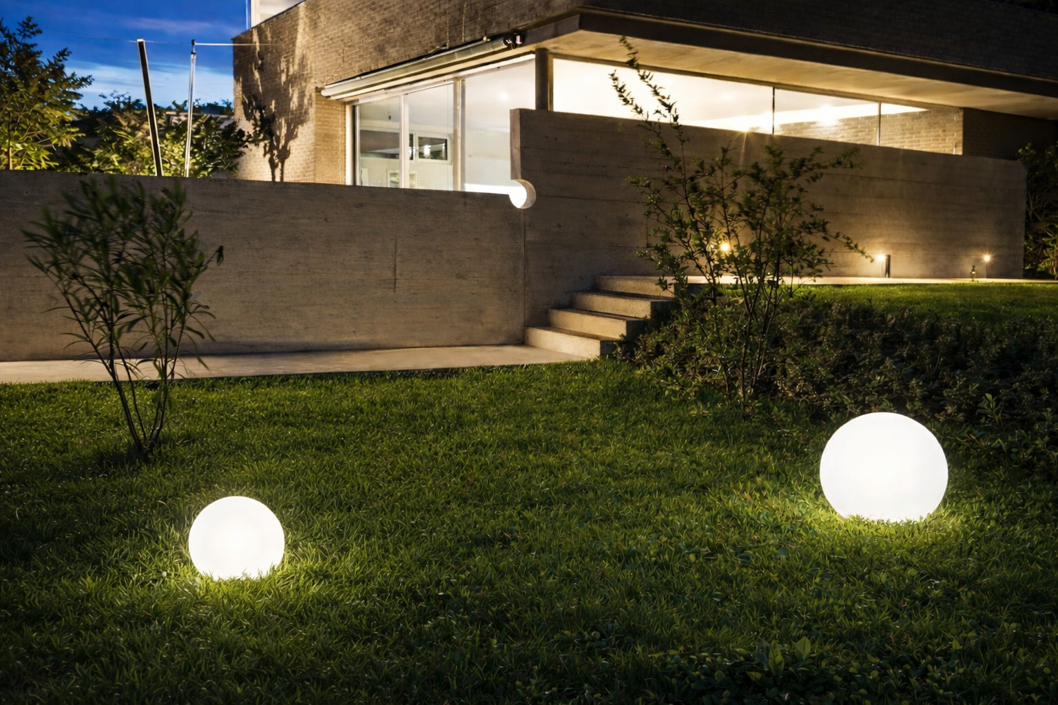 Beleuchteter moderner Garten bei Nacht mit zwei kugelförmigen Gartenleuchten auf dem Rasen vor einem modernen Haus mit großen Glasflächen.