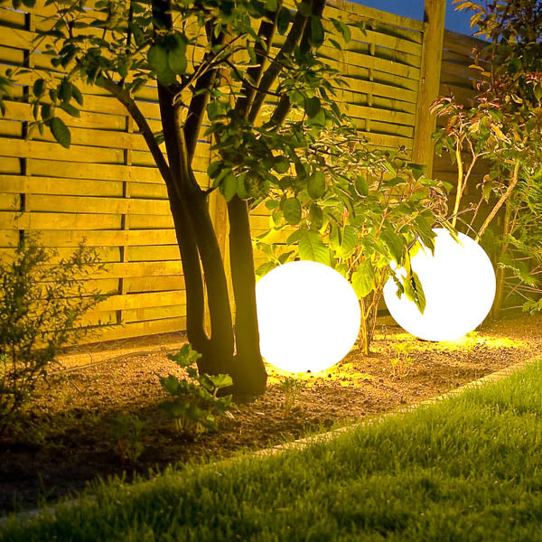 Zwei leuchtende Kugelleuchten in einem Gartenbeet neben einem Baum und Sträuchern vor einem Holzzaun, aufgenommen bei Nacht.